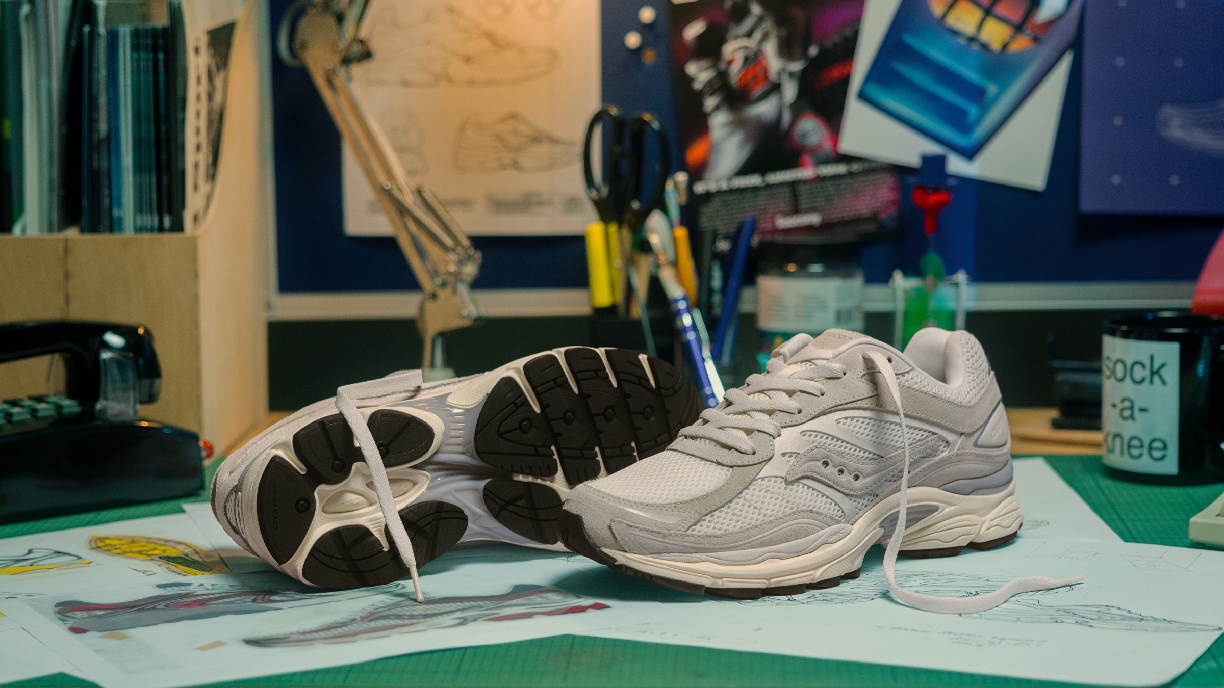 Two white and grey Saucony sneakers on a designer's desk among shoe sketches